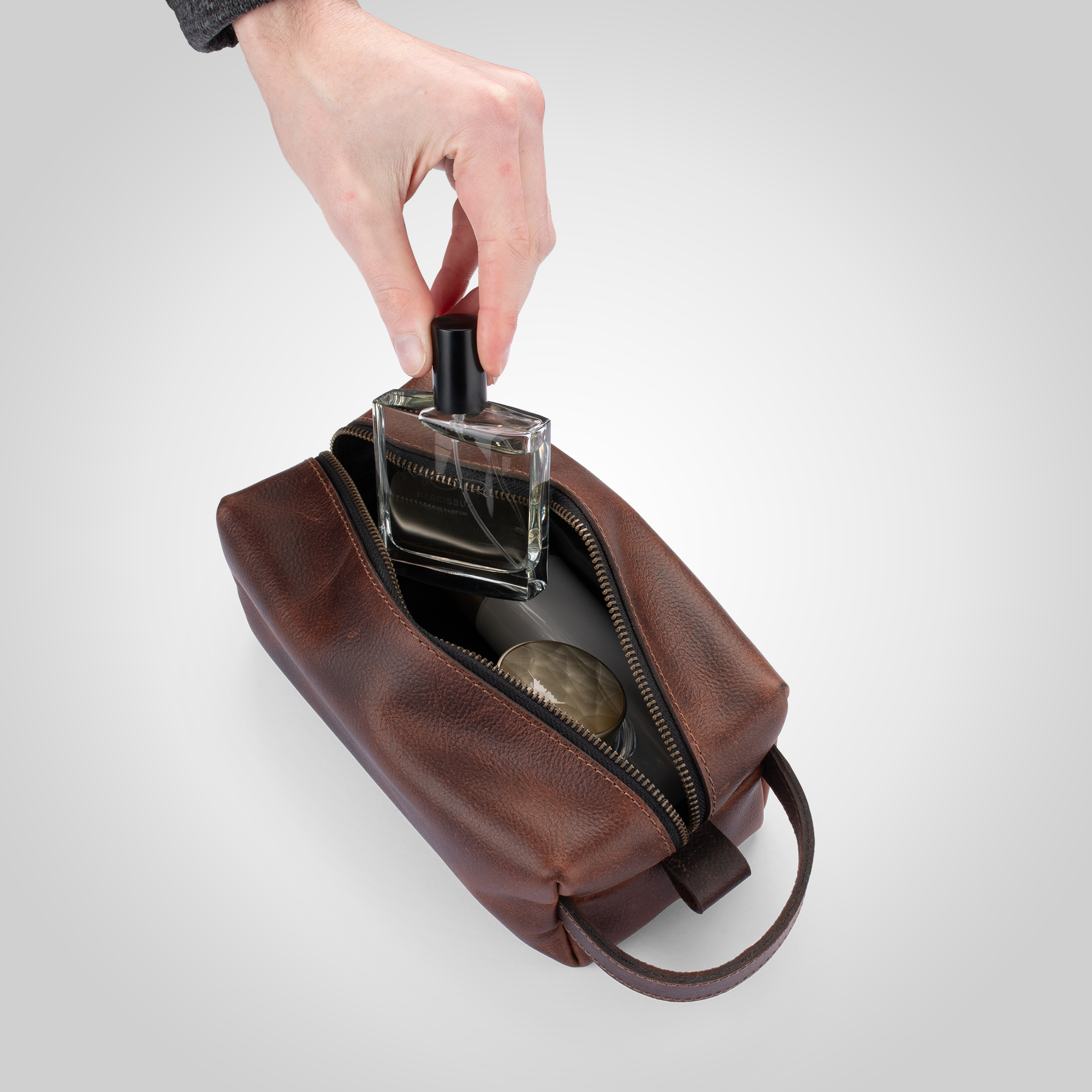 Brown leather toiletry bag with a hand placing a perfume bottle inside on a light gray background