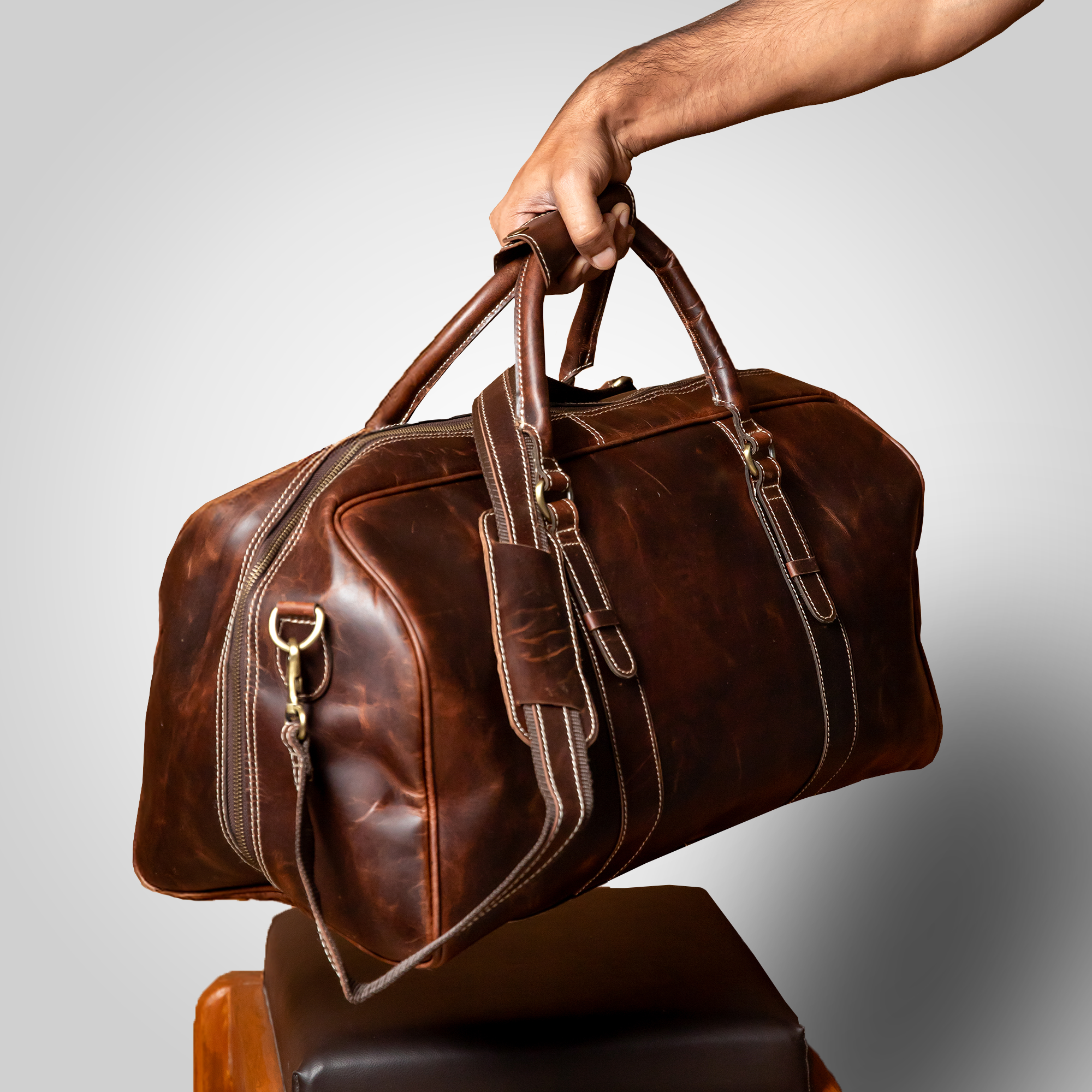 Brown leather duffel bag held by a hand against a gray background