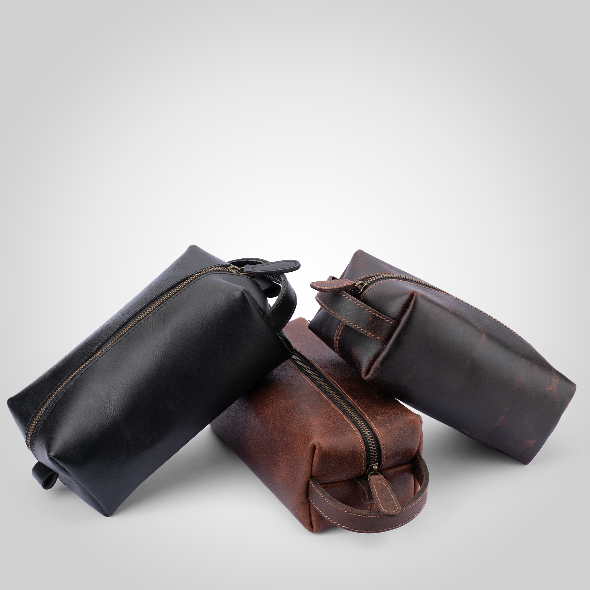 Three leather toiletry bags in black, brown, and dark brown on a light gray background.